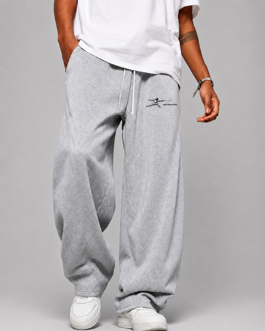 A person wearing light gray oversized sweatpants with a small black chest logo, paired with a white t-shirt and white sneakers, shown from the waist down against a light gray studio background.