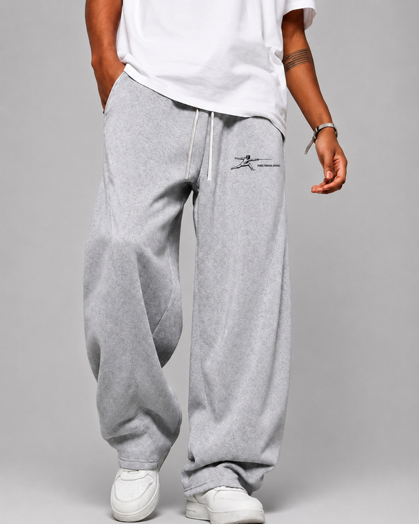 A person wearing light gray oversized sweatpants with a small black chest logo, paired with a white t-shirt and white sneakers, shown from the waist down against a light gray studio background.