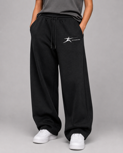 A person wearing black wide-leg sweatpants with a white graphic logo on the left thigh, paired with a dark gray top and white sneakers, photographed against a neutral gray studio background.