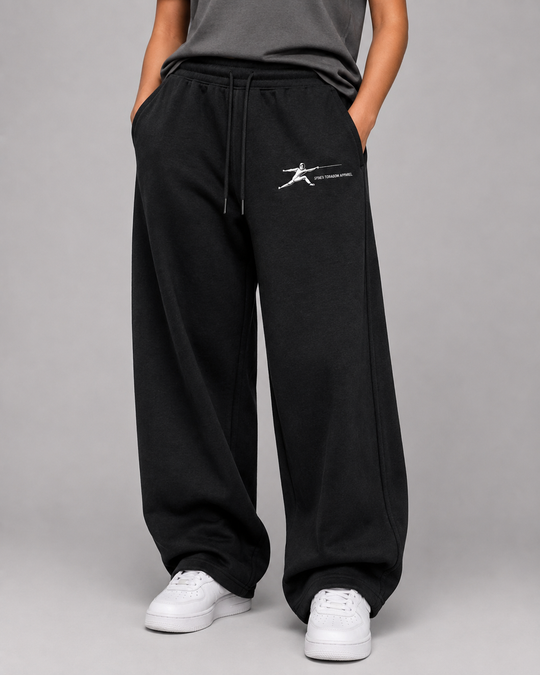 A person wearing black wide-leg sweatpants with a white graphic logo on the left thigh, paired with a dark gray top and white sneakers, photographed against a neutral gray studio background.