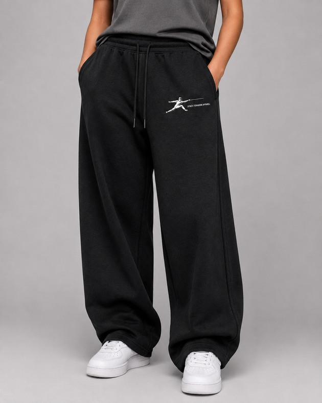 A person wearing black wide-leg sweatpants with a white graphic logo on the left thigh, paired with a dark gray top and white sneakers, photographed against a neutral gray studio background.